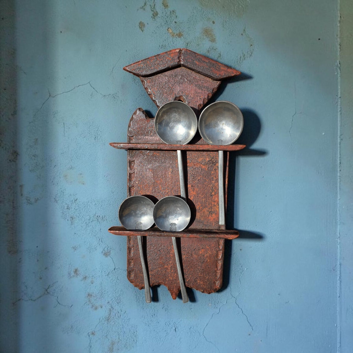 Late 17th century Low Countries painted spoon rack with gabled hood, dressed with four pewter spoons for display, hanging on a weathered blue wall.