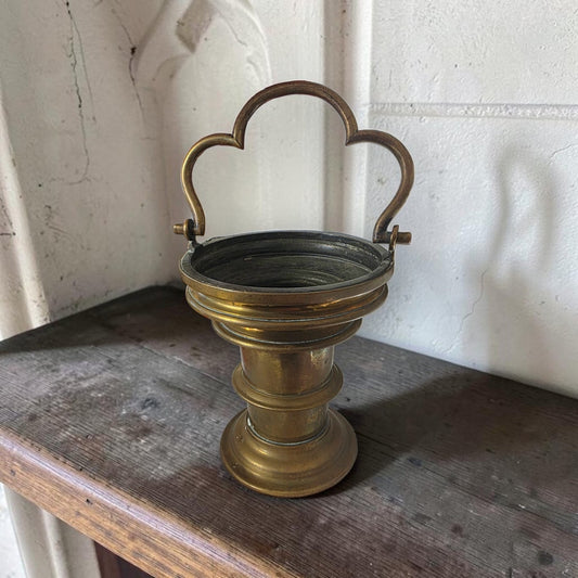 Gothic Revival brass holy water bucket with cusped trefoil handle on a weathered oak shelf before a stone Gothic arch