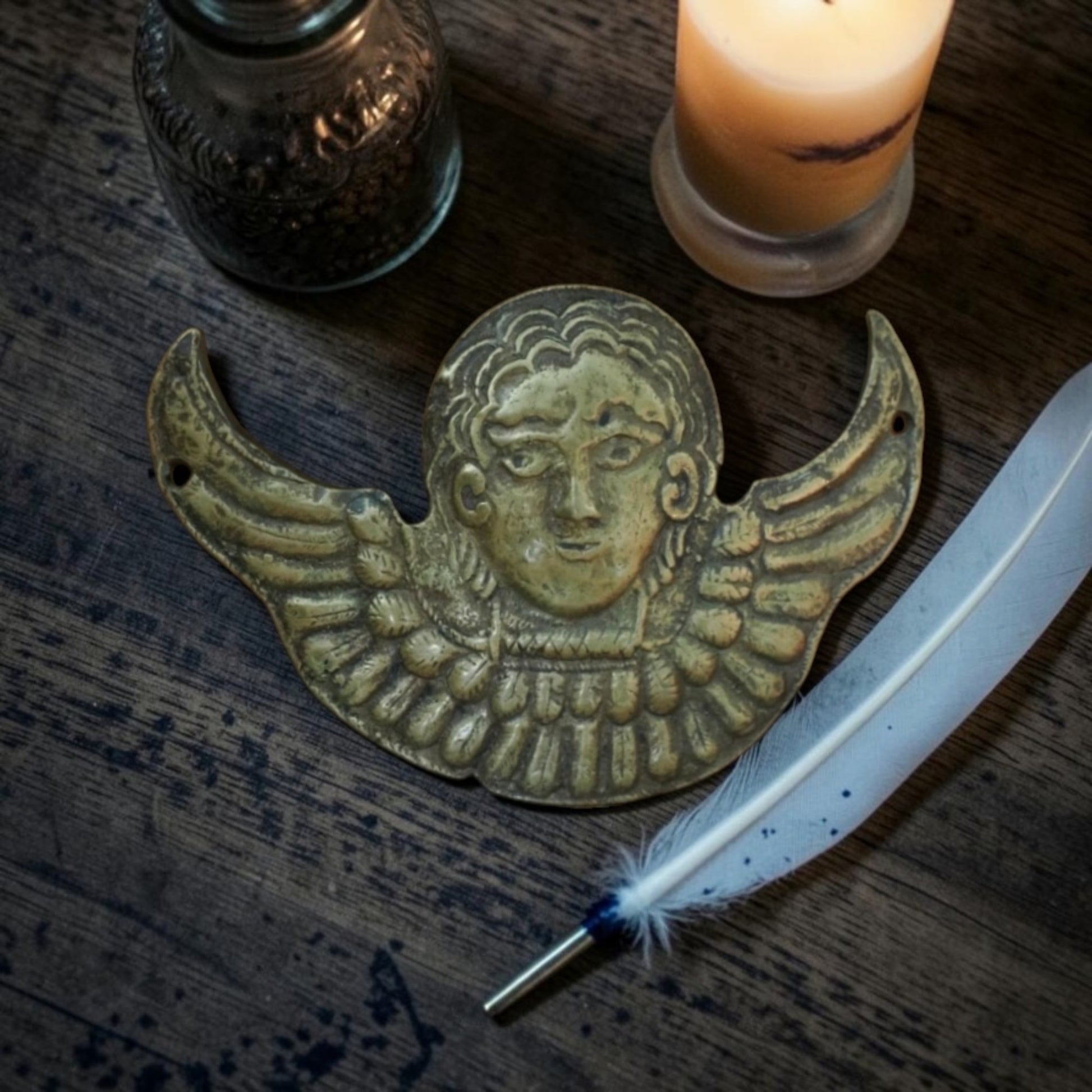  Renaissance brass winged cherub appliqué displayed on oak table beside quill and candlelight.